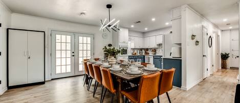 A modern kitchen with white cabinets, a dining area with a wooden table and orange chairs, and a hallway with a white door and a plant.