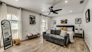 Ein modernes Schlafzimmer mit grauem Sofa, einem Bett mit dunklem Kopfteil, einem Nachttisch und einem Deckenventilator.