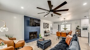 Ein modernes Wohnzimmer mit Kamin, Deckenventilator und Essbereich, im Hintergrund eine Küche.