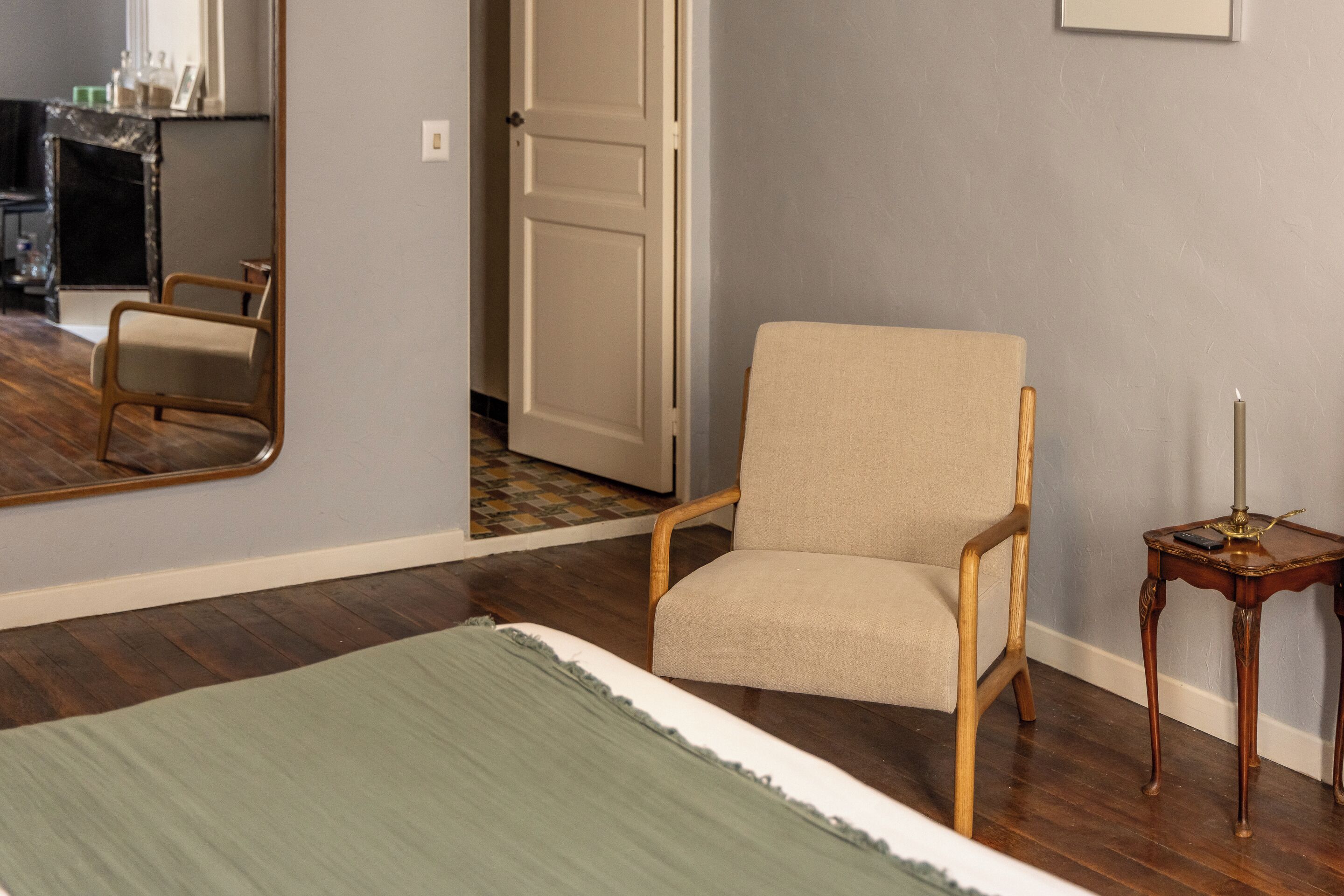 Una habitación con cama, una silla, una mesita de noche con lámpara y un espejo en la pared.