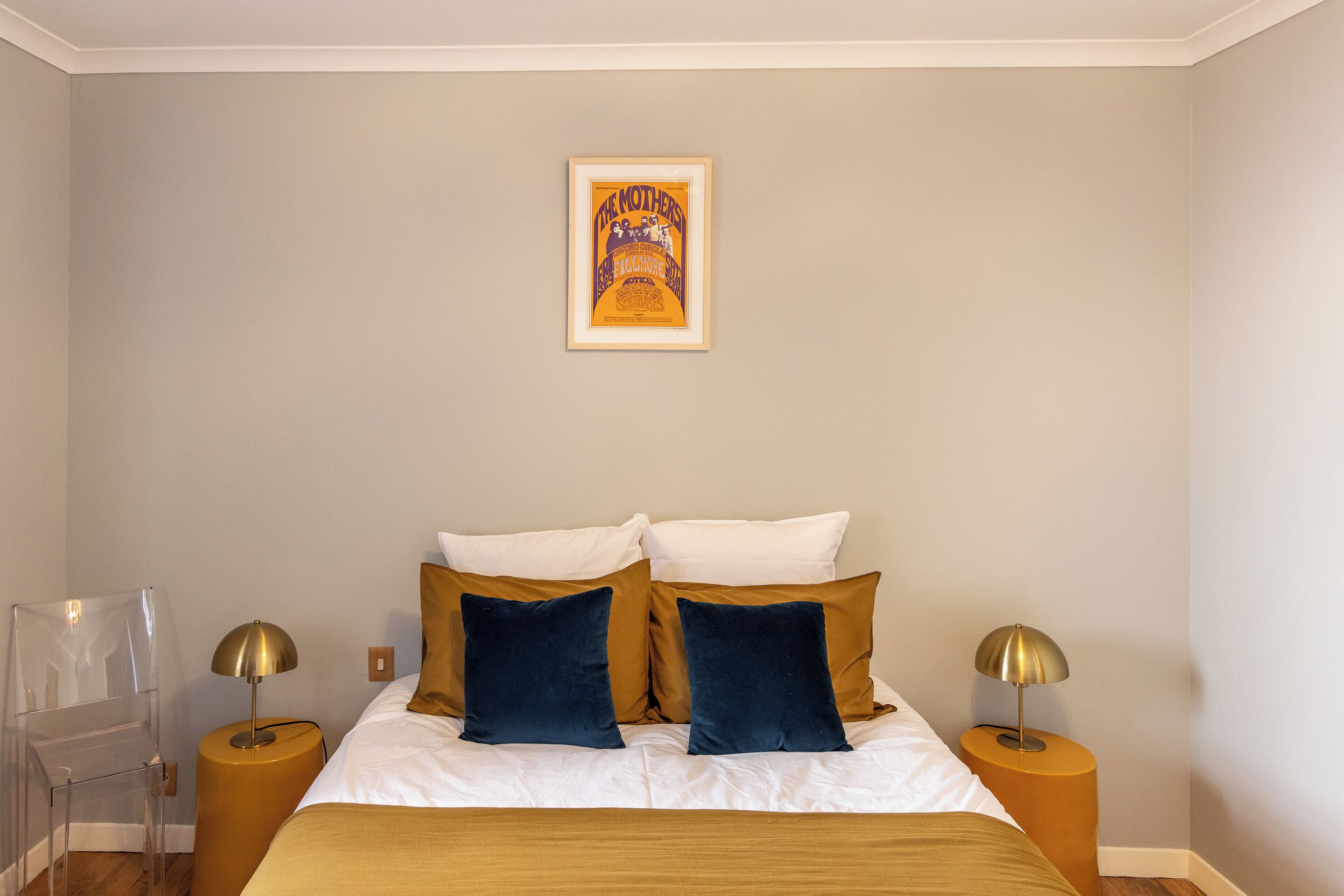 A neatly arranged bedroom with a bed, two nightstands, a clear chair, and a framed poster on the wall.