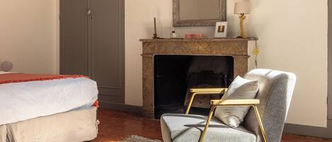 A bedroom with a bed, a chair, a fireplace, and a mirror.