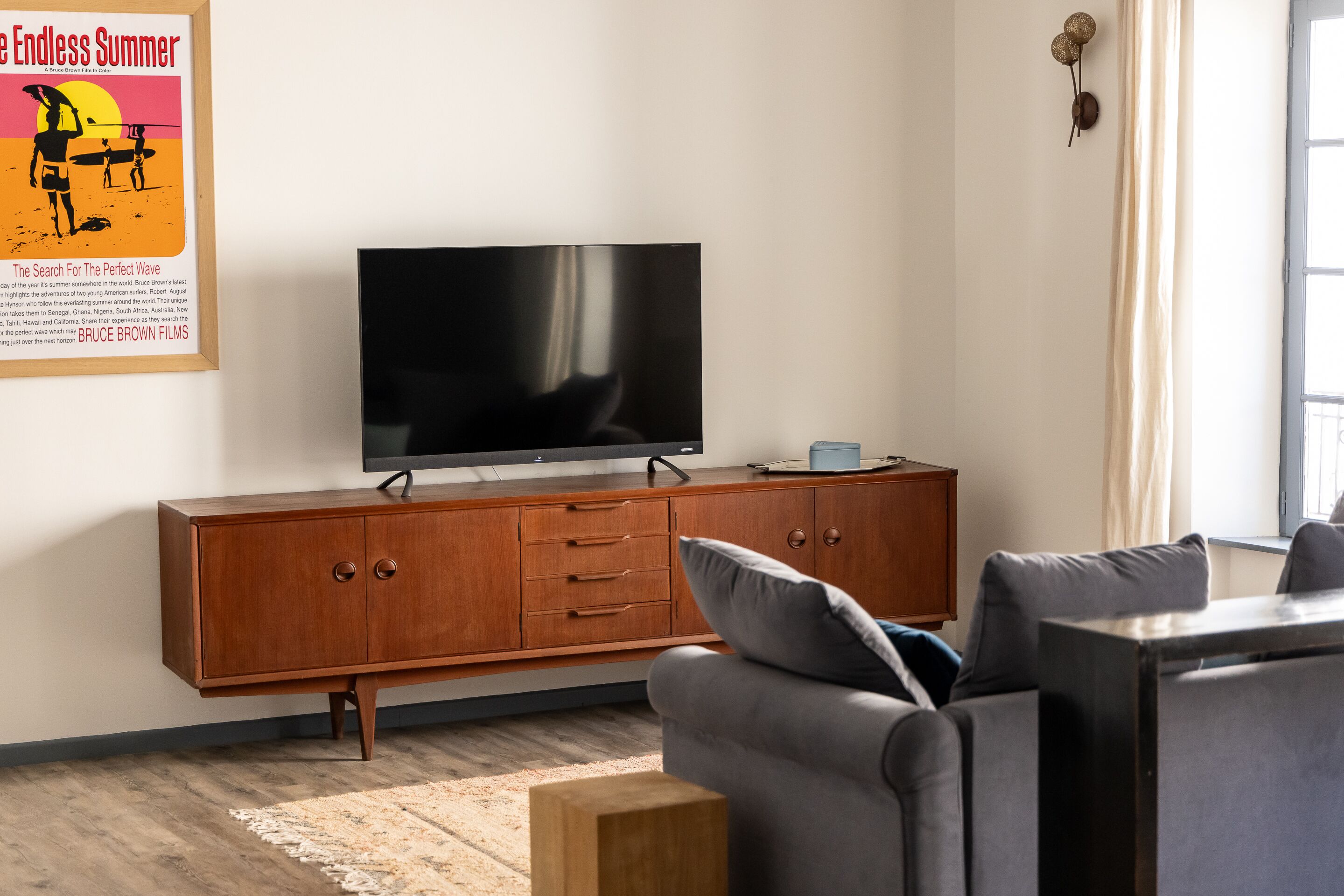 Ein Wohnzimmer mit Flachbildfernseher auf einem Holzkabinett, einem grauen Sofa und einem kleinen Holzschemel.