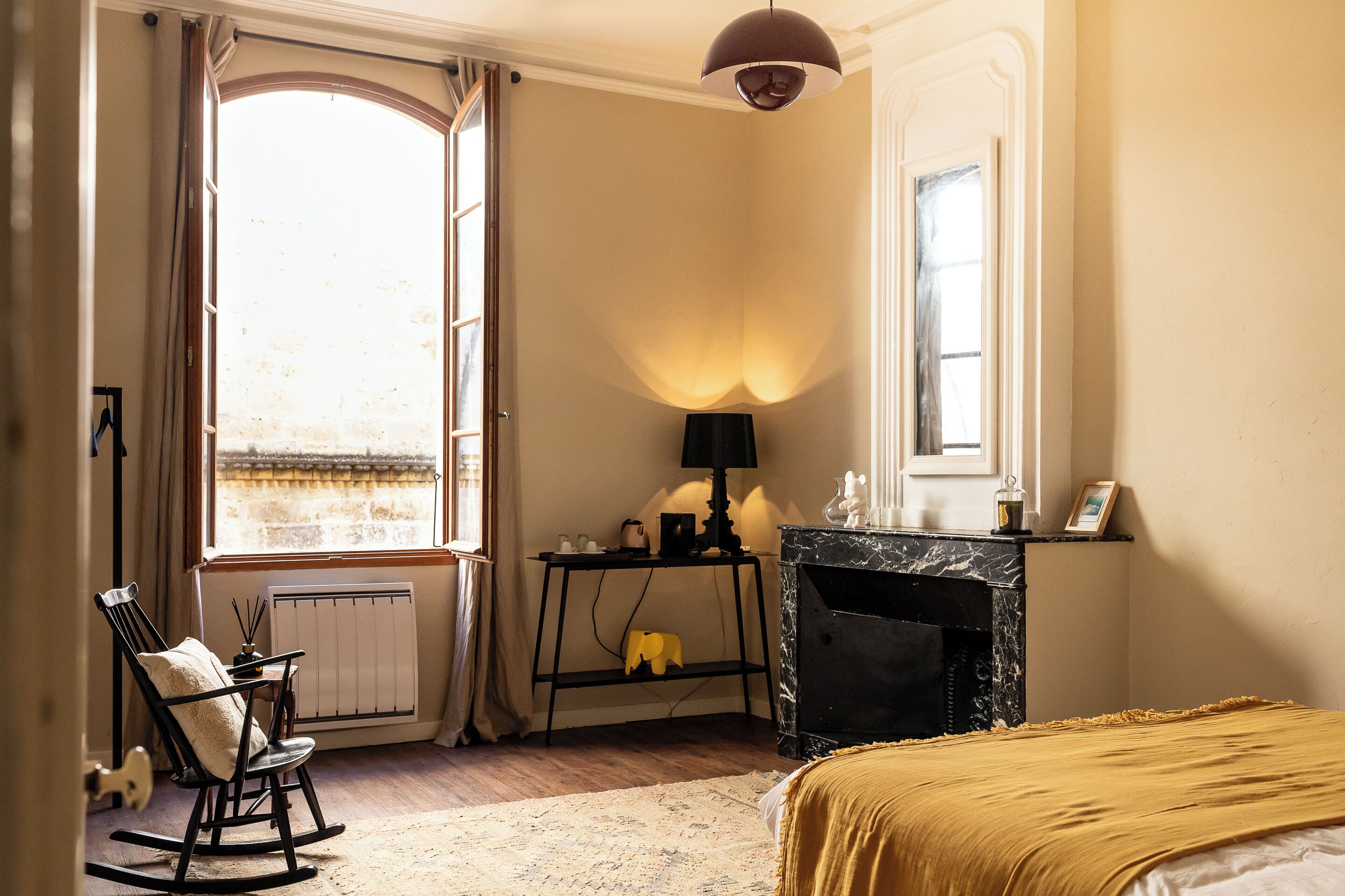 Una habitación con chimenea, un sillón mecedor, una mesa con lámpara y un ventanal grande.