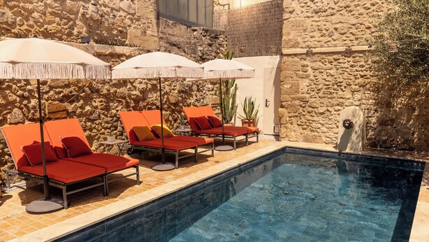 Zona junto a la piscina con sillones de descanso y sombrillas, un muro de piedra y una planta en maceta.