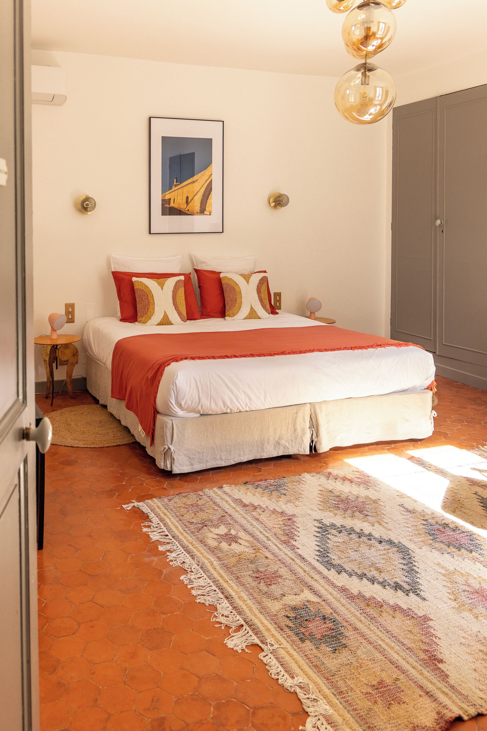 A bedroom with a large bed, a patterned rug, a framed picture on the wall, and a door to the left.