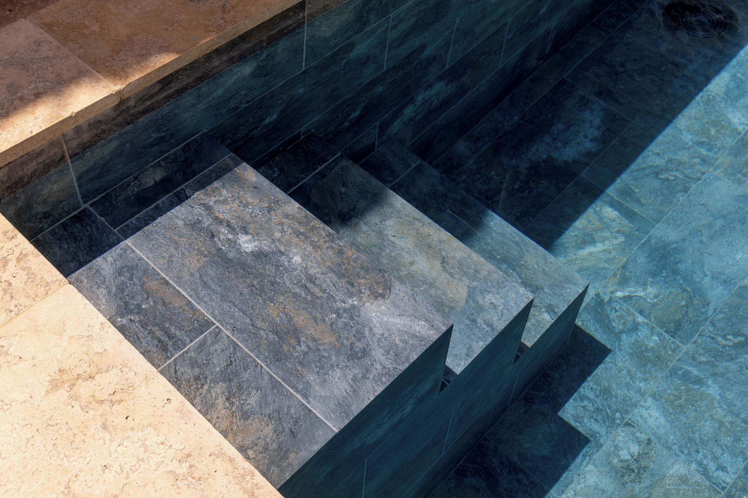 Une piscine dont le fond est recouvert de carreaux clairs et foncés, dont l’eau est limpide et qui est bordée d’une margelle en pierre.