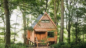 A wooden cabin in a forest with a triangular roof, a wooden deck, and a window. - Luxury Treehouse with amazing views in 100 acres of secluded private Land. (Hereford)