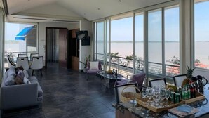 Ein geräumiges Zimmer mit großem Fenster und Blick auf ein Gewässer, eine Sitzecke mit Esstisch und ein modernes Interieur.