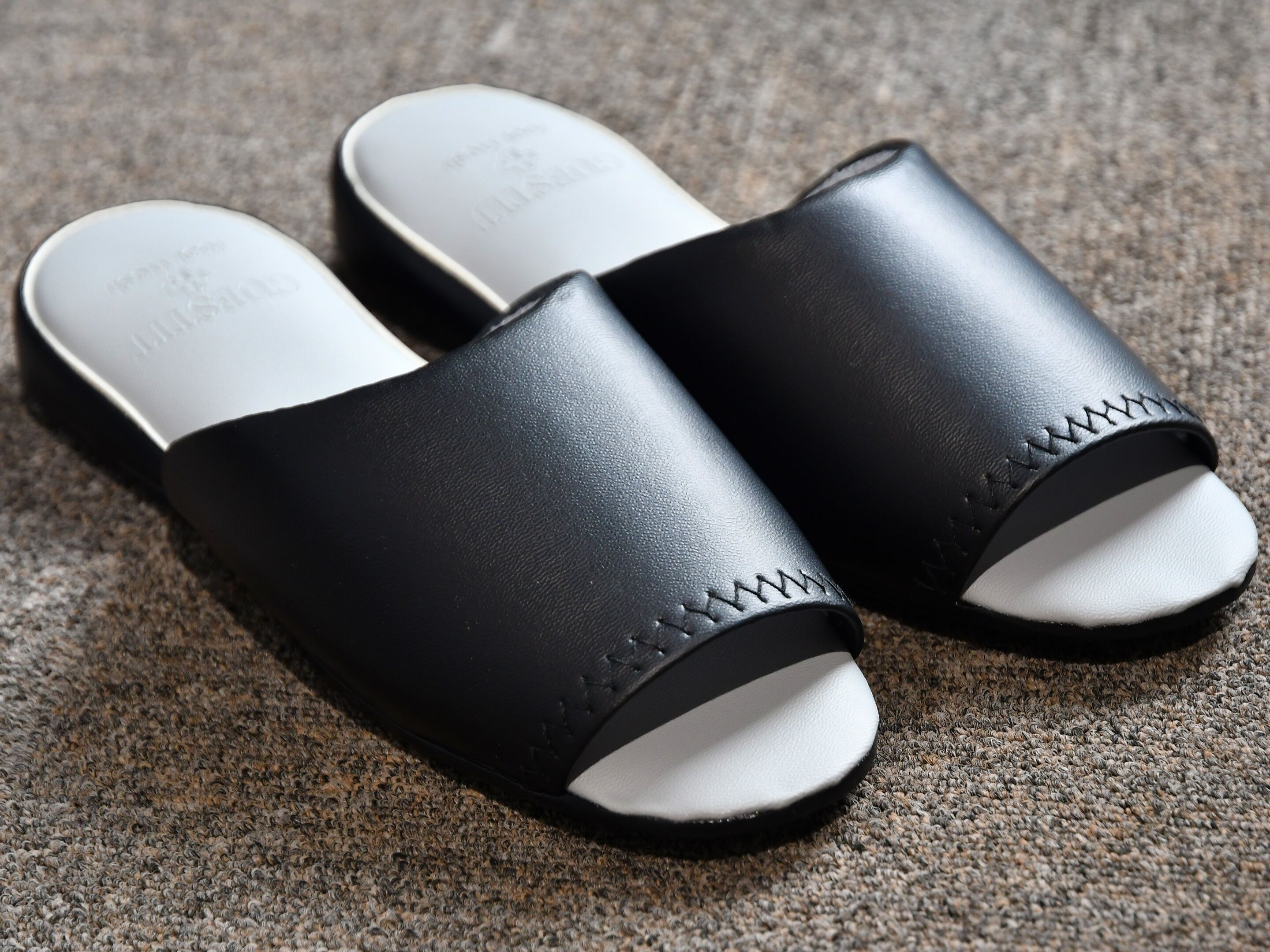 Ett par svarta sandaler med vita sulor och ett sicksackmönster på sidan, placerade på en strukturerad yta.