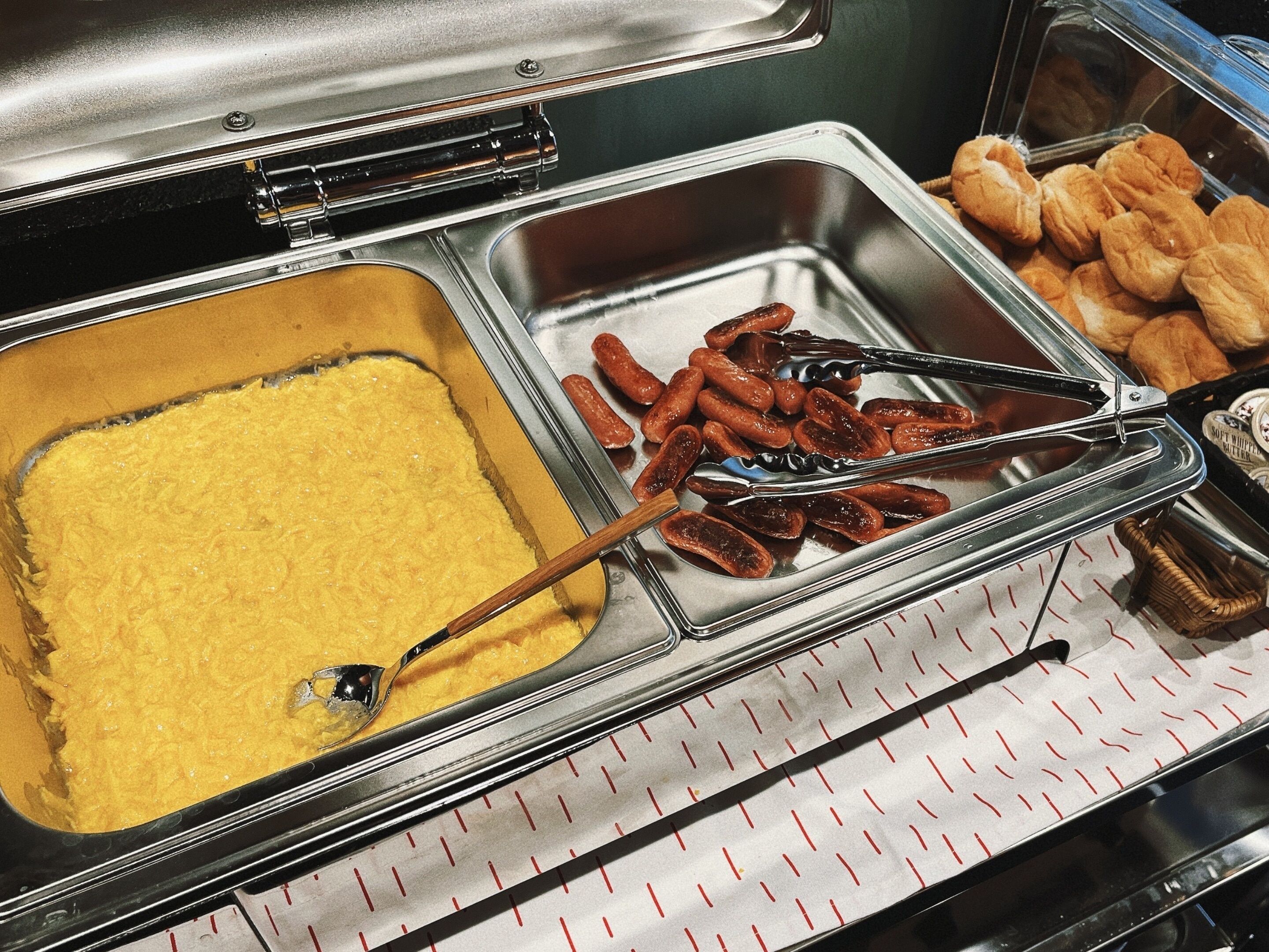 A buffet with a variety of food items including a tray of yellow custard, sausages, and pastries.