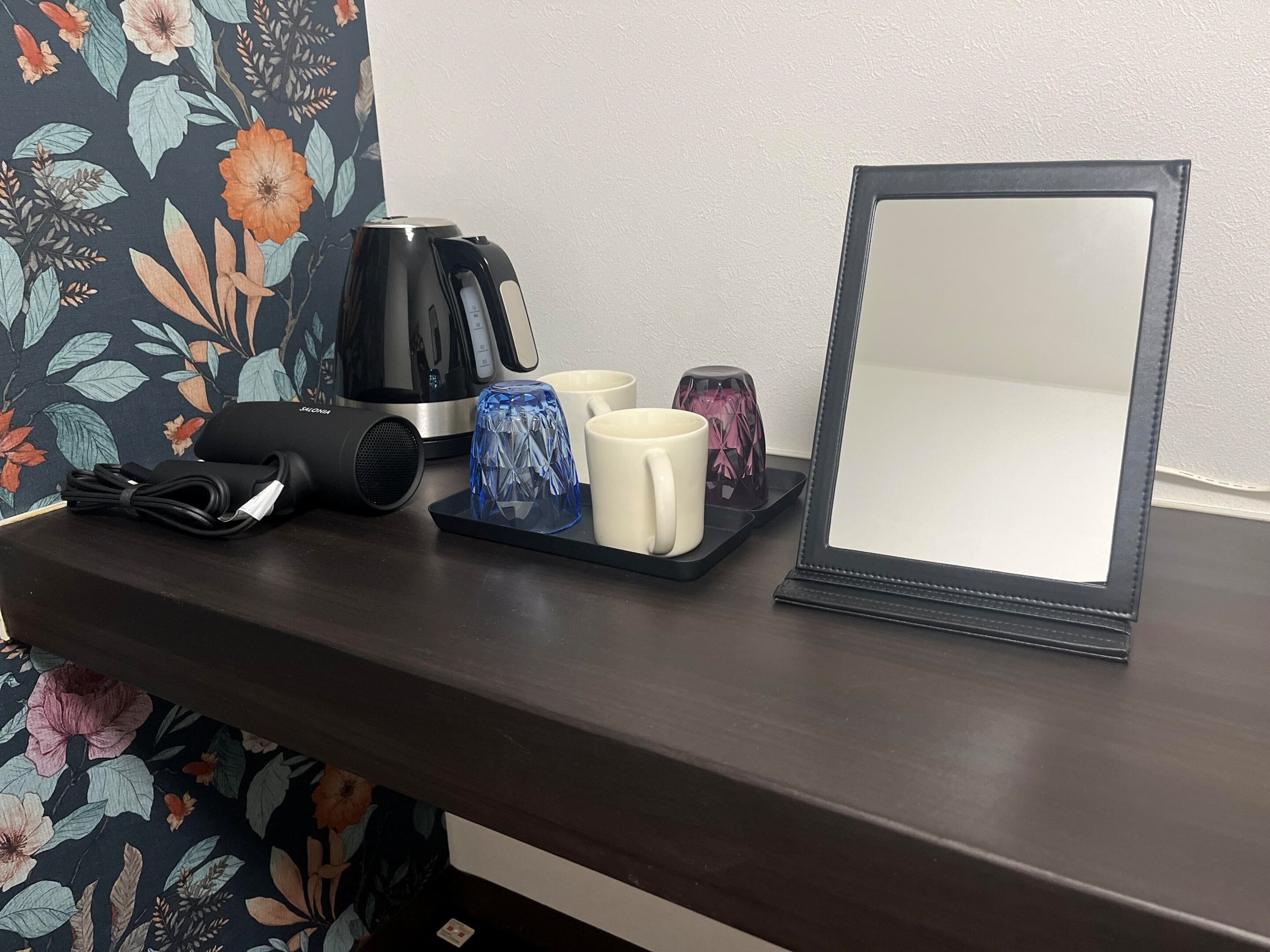 A dark wooden table with a black kettle, a blue glass, a white mug, a pink glass, and a black speaker.