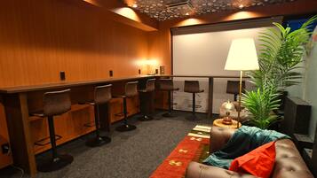 A conference room with a long table, multiple bar stools, a sofa, a projector screen, and a large plant.