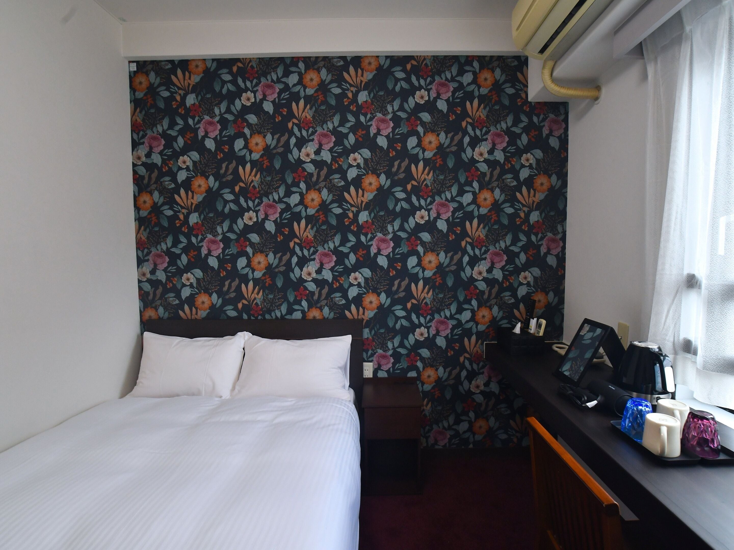 A hotel room with a bed, a desk with a coffee maker, and a floral-patterned wall.