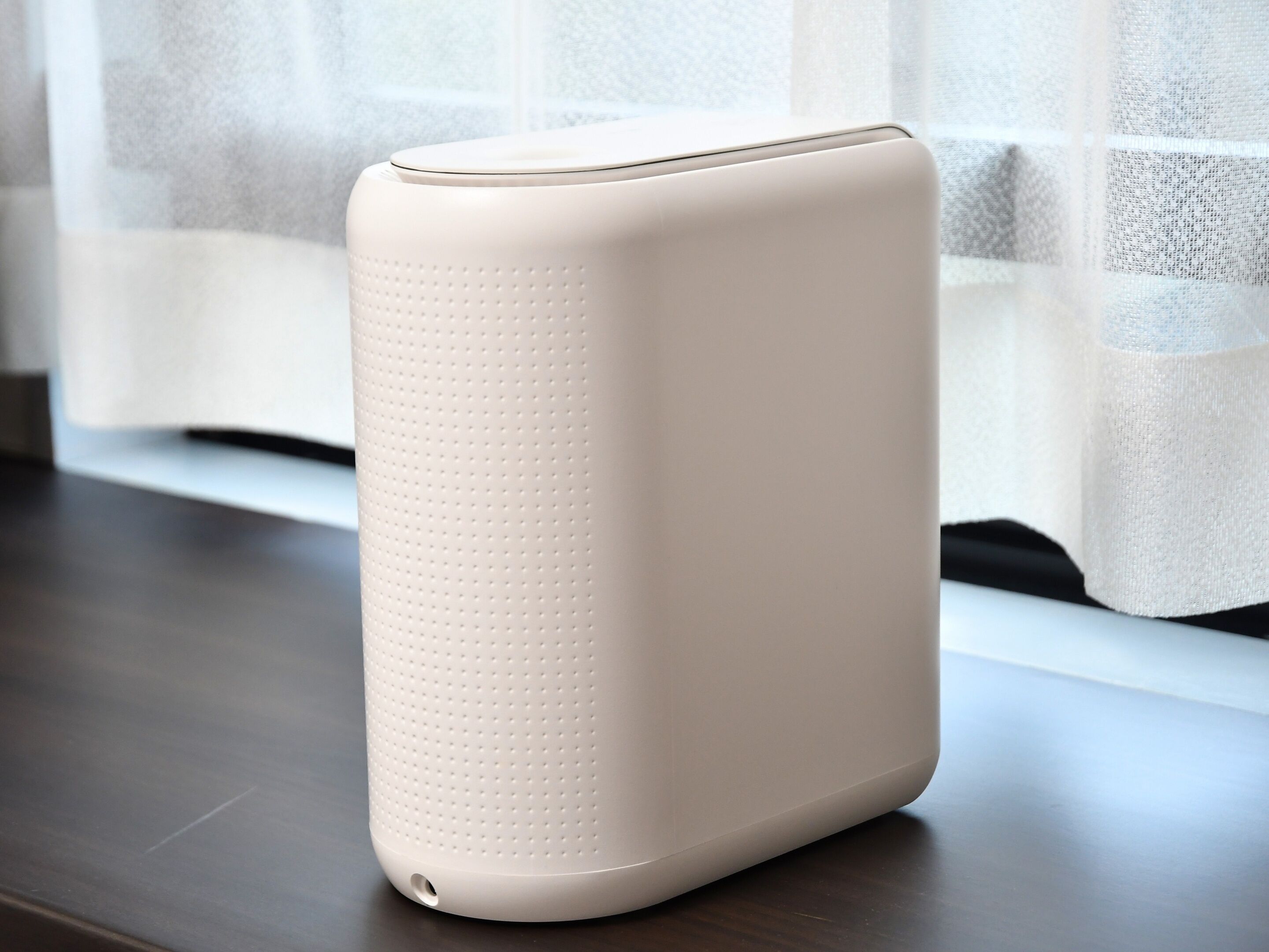 Un haut-parleur blanc, rectangulaire et portable, doté d’une surface perforée, placé sur une surface en bois près d’une fenêtre ornée de rideaux légers.