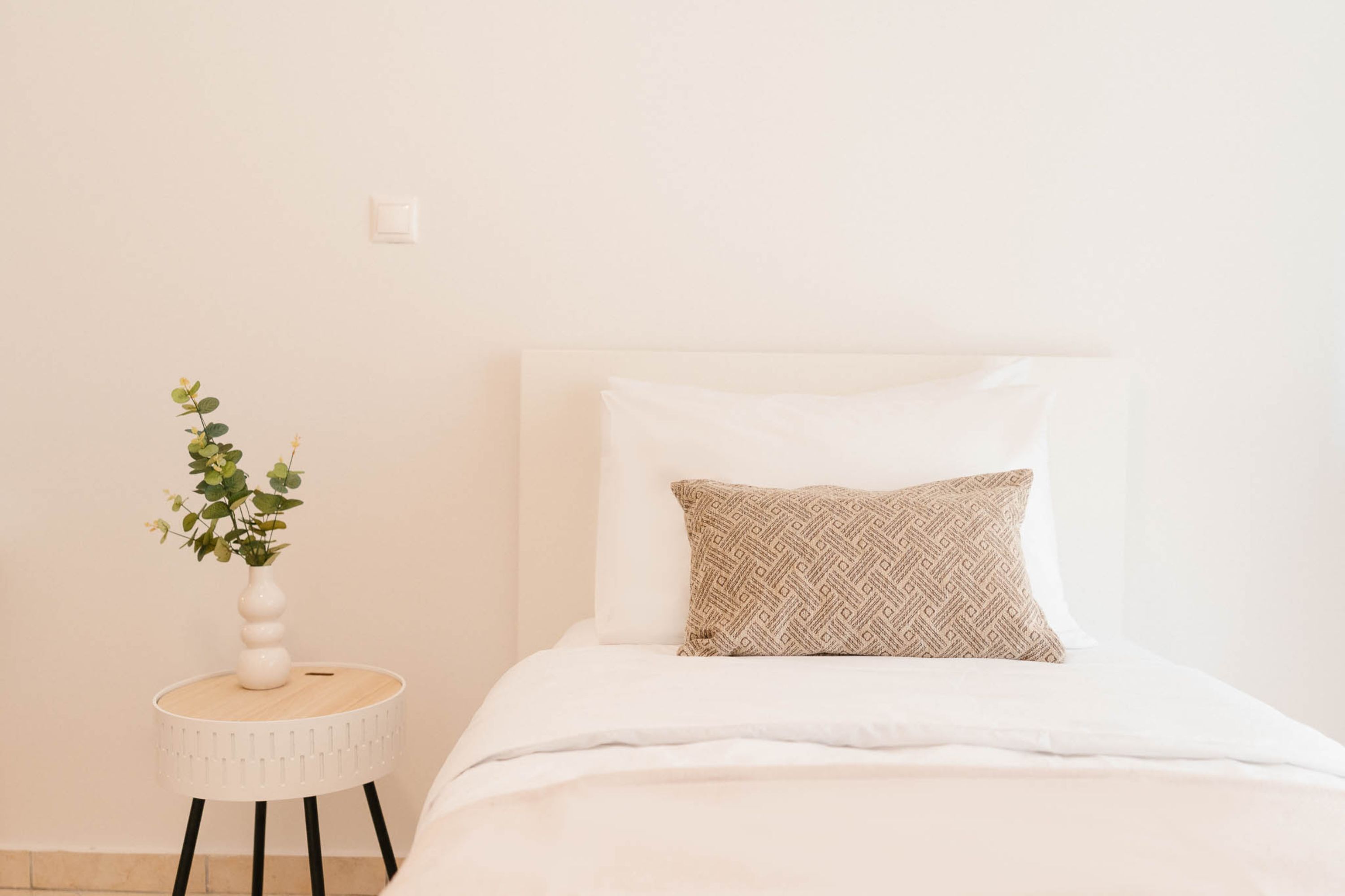 A neatly made bed with a patterned pillow and a white vase with green plants on a small round side table.
