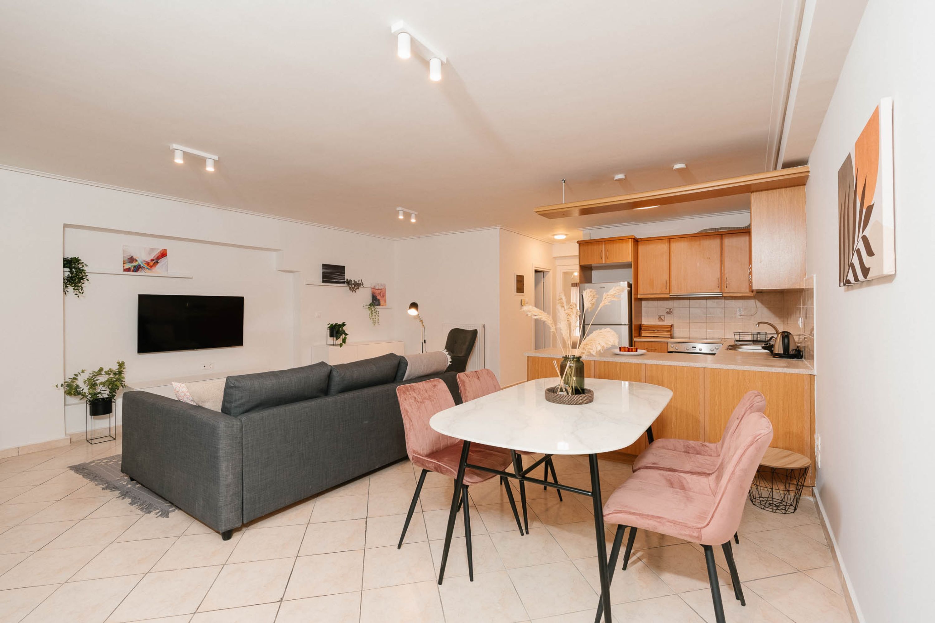 A modern living room with a grey sofa, a dining table with pink chairs, and a kitchen area with wooden cabinets.