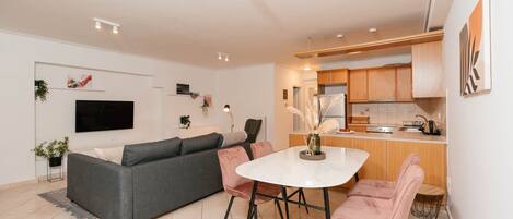 A modern living room with a grey sofa, a dining table with pink chairs, and a kitchen area with wooden cabinets.