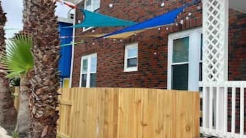 Une maison à deux étages en briques, avec une clôture en bois, une voile d’ombrage bleue et un palmier.