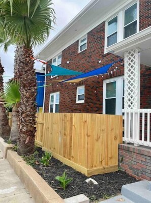 Una casa de ladrillo de dos pisos, con cerca de madera, una vela de sombra azul y una palmera.
