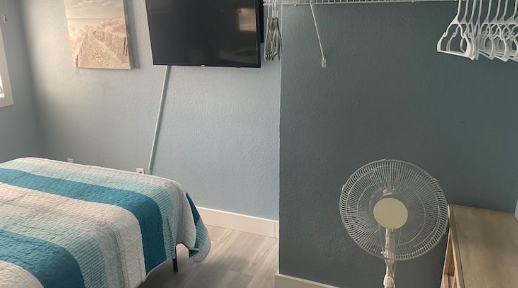A bedroom with a bed, a wall-mounted TV, and a standing fan.