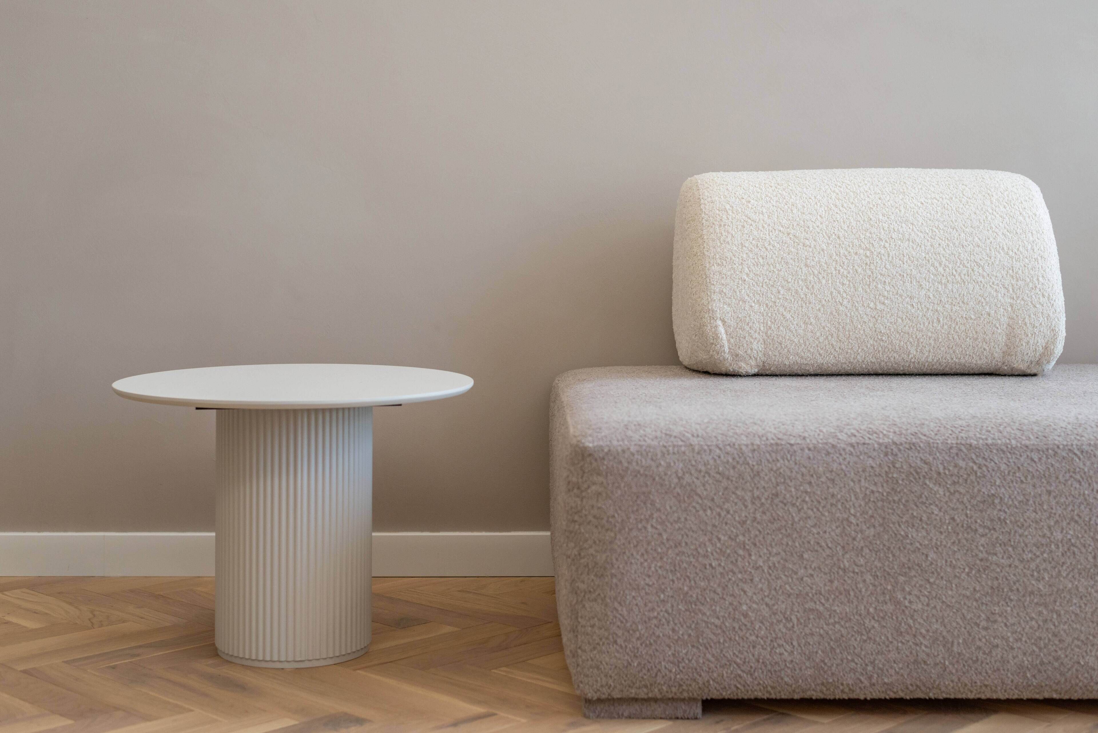 Een ronde witte tafel met een geribbeld onderstel en een grijze gestoffeerde bank met een kussen, voor een effen muur en houten vloer.
