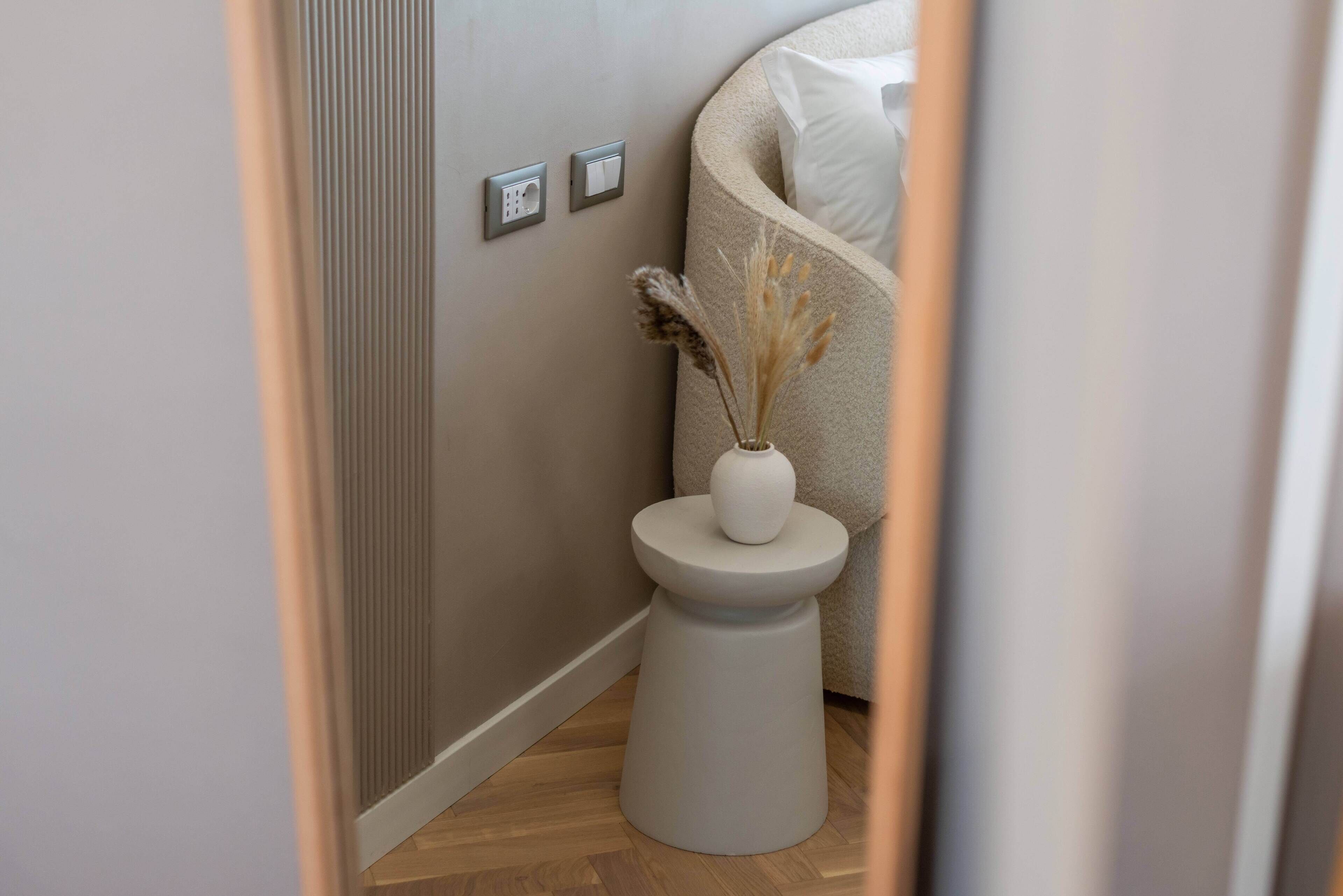 Een smalle gang met een witte bijzettafel waarop een vaas met gedroogde planten staat, een beige bank met een wit kussen, en een in de muur bevestigde lichtschakelaar.