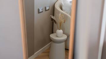 Een smalle gang met een witte bijzettafel waarop een vaas met gedroogde planten staat, een beige bank met een wit kussen, en een in de muur bevestigde lichtschakelaar.