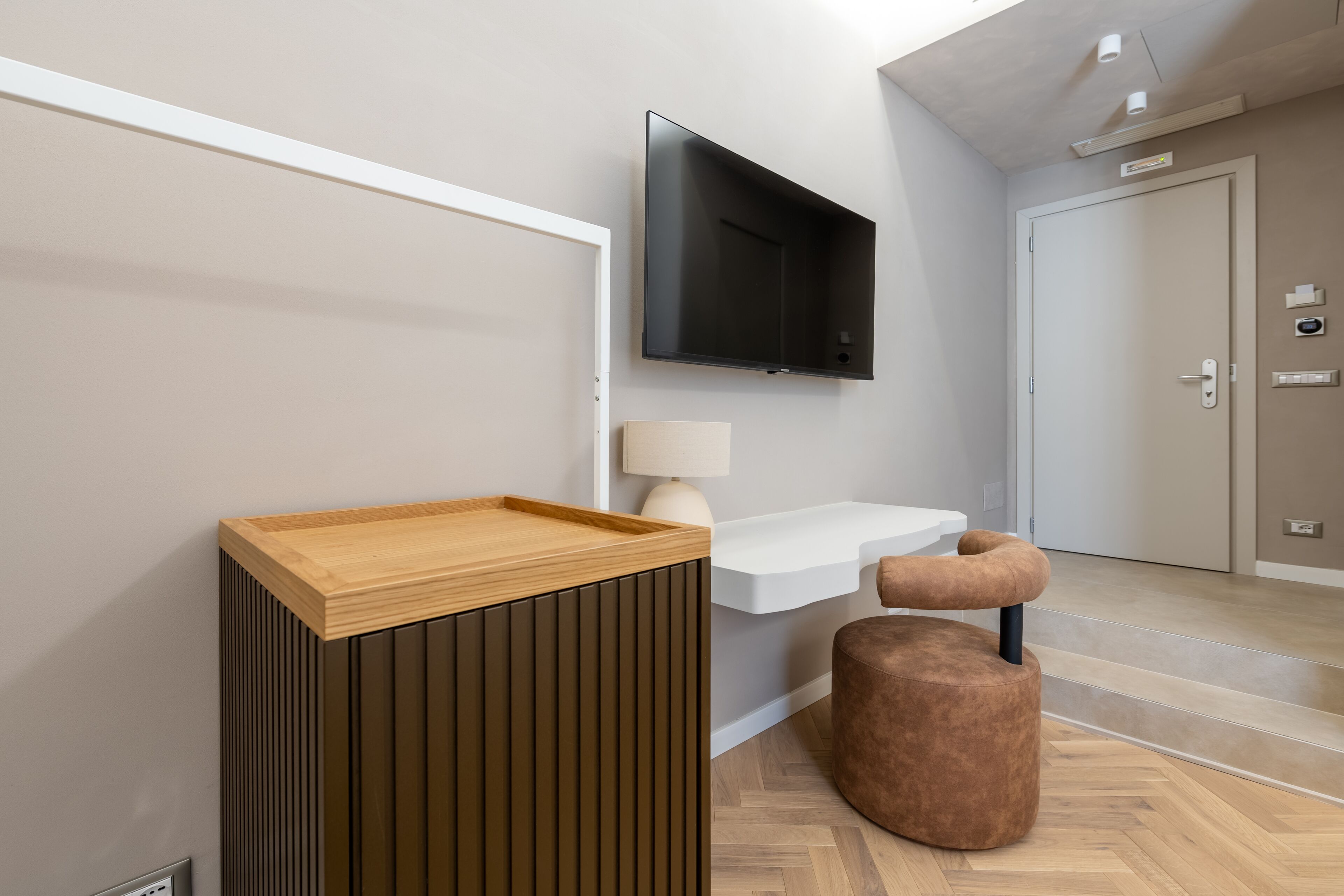 A modern hotel room with a wooden console, a white bedside table, a brown ottoman, and a mounted TV.