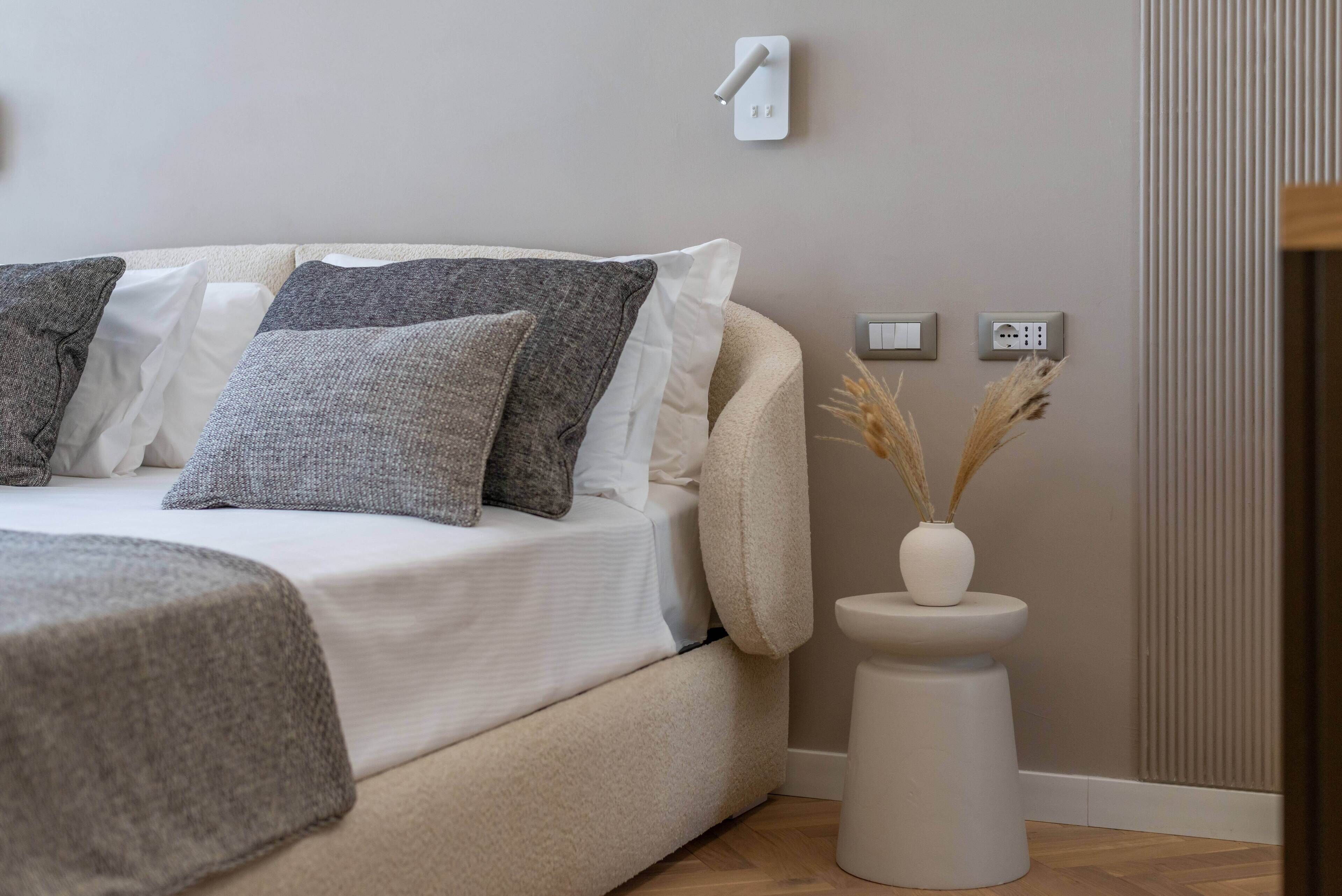 A modern bedroom with a bed, a bedside table with a vase, and a wall-mounted light switch.