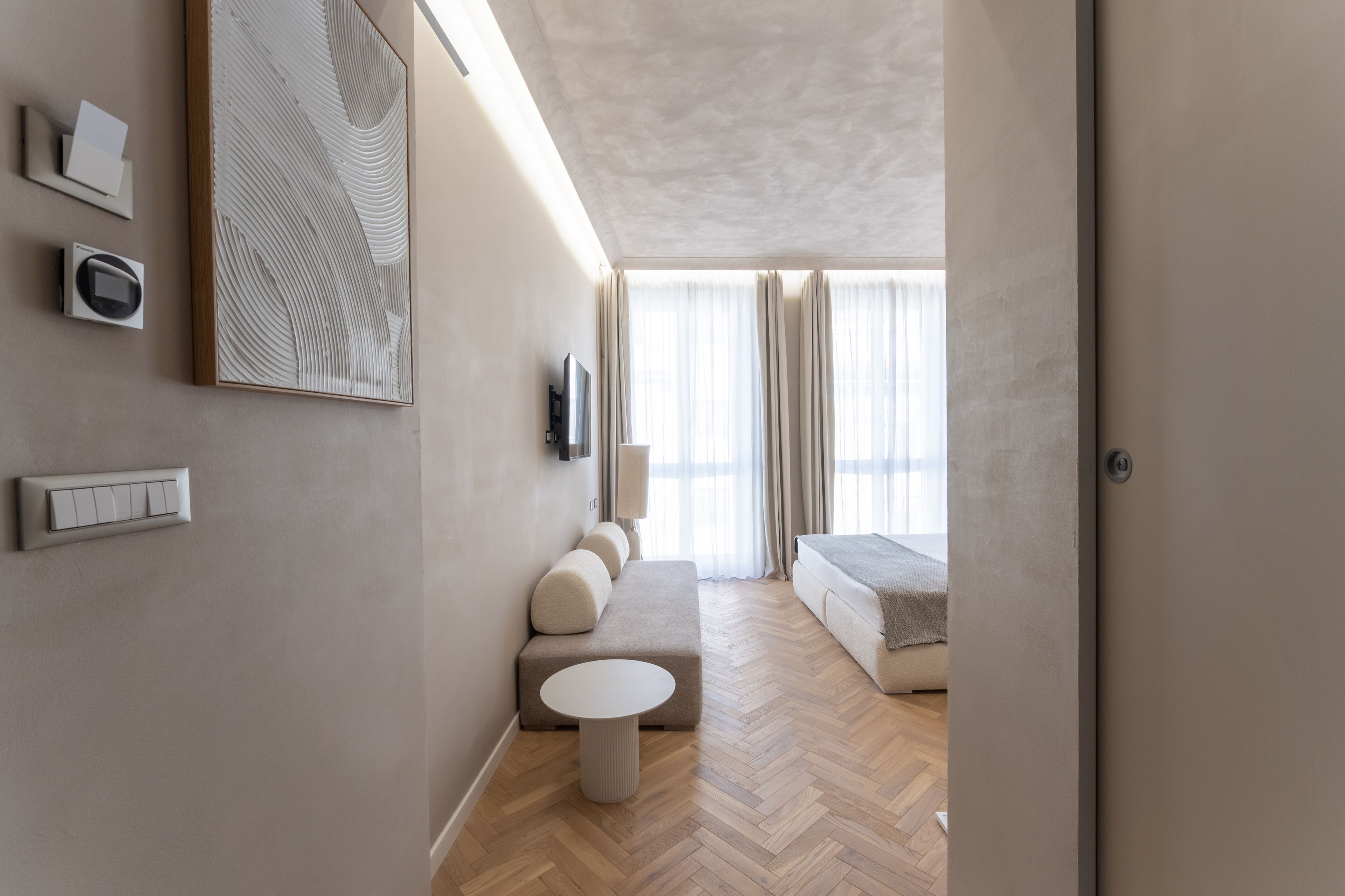 A modern hotel room with a herringbone wood floor, a round white table, a beige sofa, and a large abstract painting on the wall.