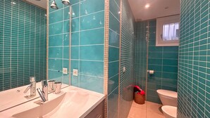 Un baño con azulejos color turquesa, un lavabo con dos grifos, una zona de ducha y un inodoro.