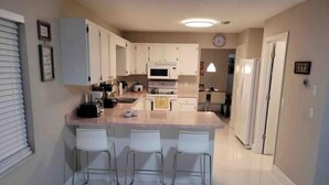 A modern kitchen with white cabinets, a central island with bar stools, a microwave, and a refrigerator. - Beautiful Relaxed Sebring Home. (Sebring)