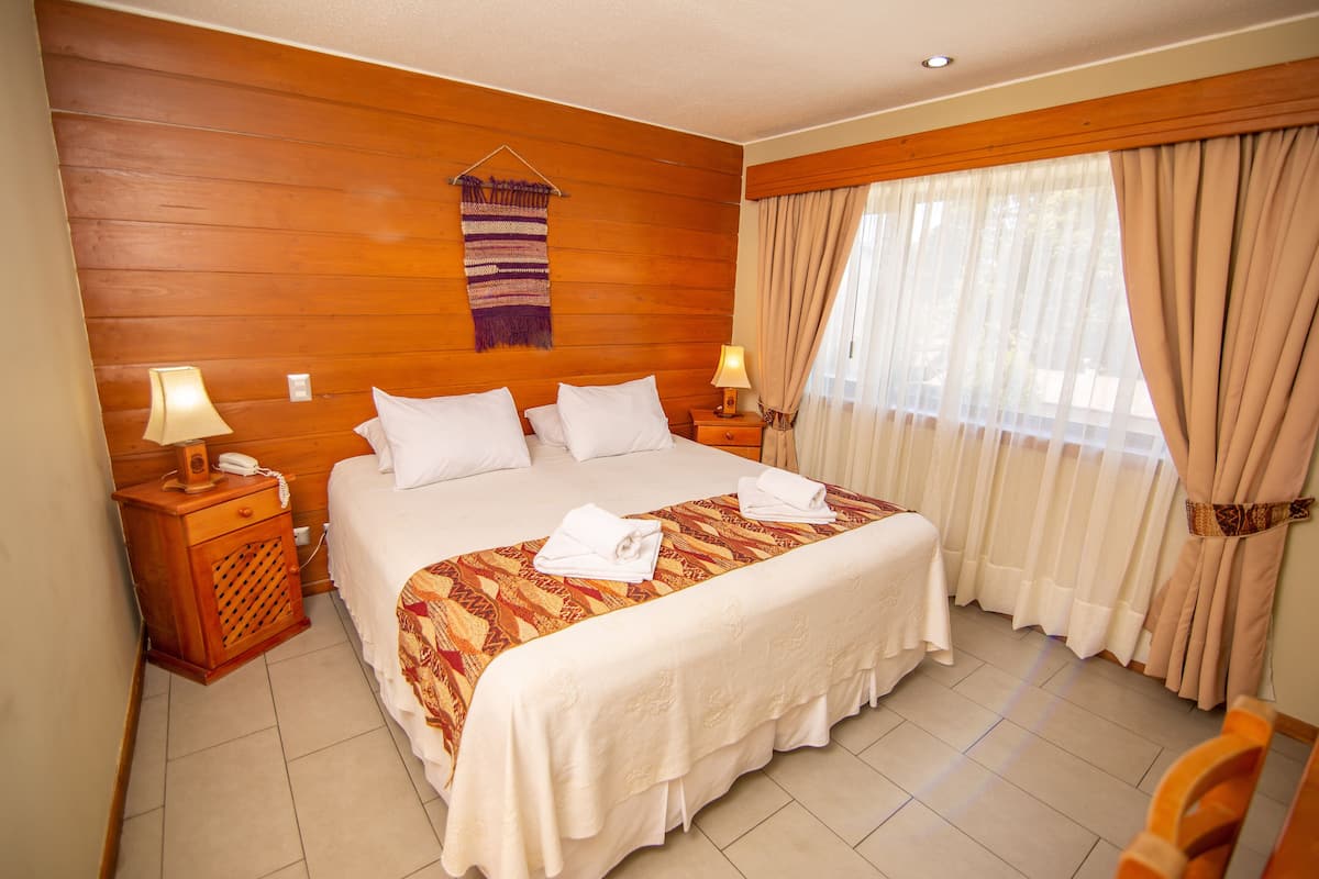 A hotel room with a large bed, wooden headboard, two bedside tables with lamps, a hanging tapestry, and a large window with curtains.