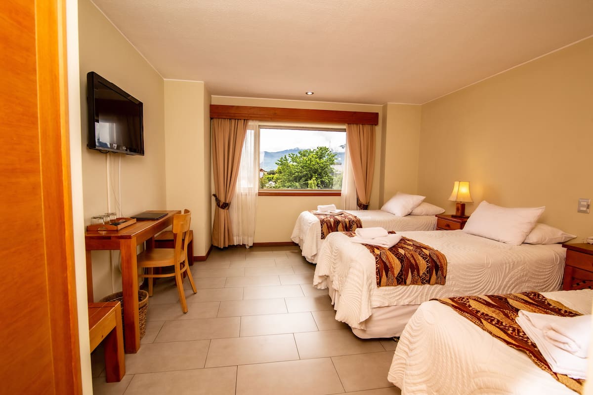 A hotel room with two beds, a desk, a chair, a TV mounted on the wall, and a window with a view of greenery and mountains.
