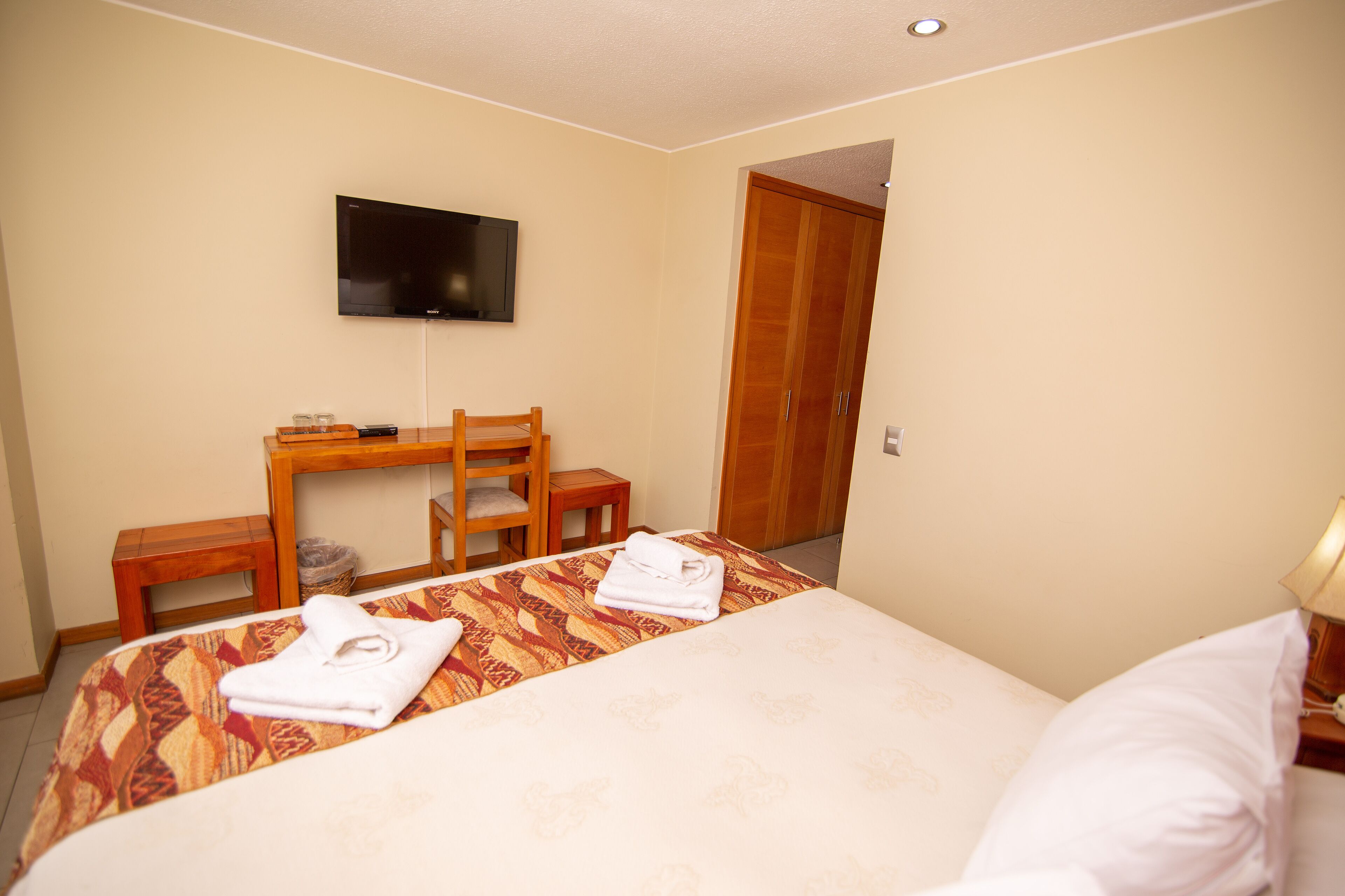 A hotel room with a bed, a TV mounted on the wall, a wooden desk, and a wardrobe.