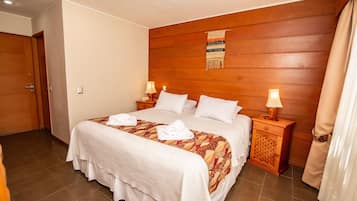 Una habitación de hotel con una cama grande, cabecera de madera, dos mesitas de noche con lámparas y una puerta.