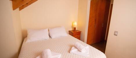A neatly made bed with white linens and two towels, a wooden headboard, a bedside lamp, and a wooden wardrobe.