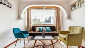 En stue med sofa, to stole, sofabord og udsigt til bylandskabet gennem et stort vindue.
