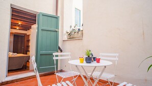 Een klein buitgedeelte met een tafel en stoelen, een groene deur en een bed dat door een open deur zichtbaar is.