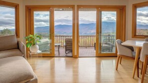 Una habitación con piso de madera, un sofá, una mesa de comedor con sillas y un balcón con vista a las montañas.