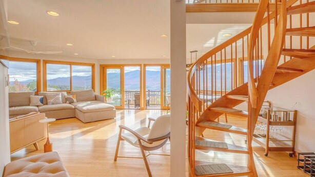 Amplia sala de estar con escalera de madera, ventanas grandes y vista a las montañas.