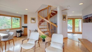 Un salón amplio con una escalera de madera, dos sillones blancos, una mesita con una planta y grandes ventanales que ofrecen vistas al exterior.