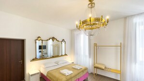 Una habitación de hotel con cama, un espejo grande, una araña, un armario y una ventana con cortinas.
