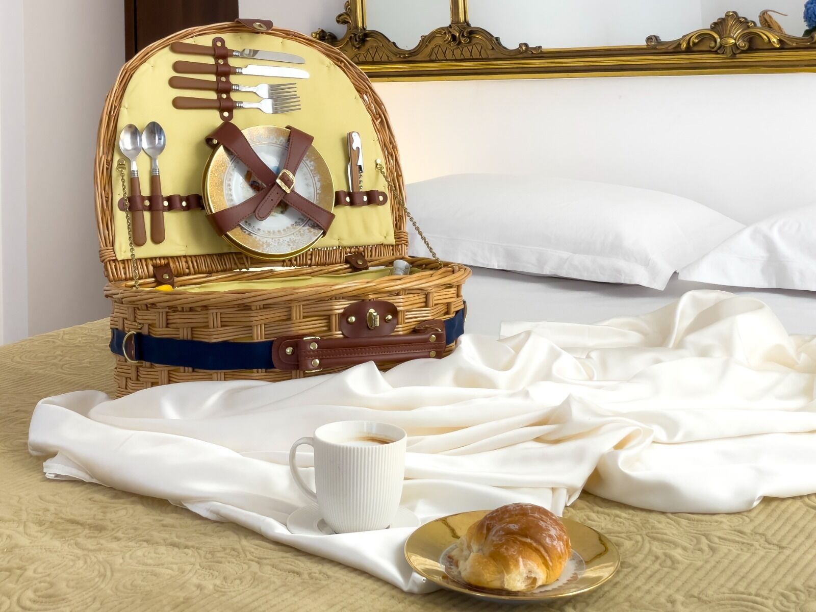 Cesto da picnic in vimini con posate, una bussola e un piatto con un croissant su un letto con lenzuola bianche.