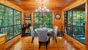 A wooden cabin interior with a round dining table, four upholstered chairs, a chandelier, and large windows offering a view of the forest. - Timber Nest | Stay Amongst the Trees (Blue Ridge)