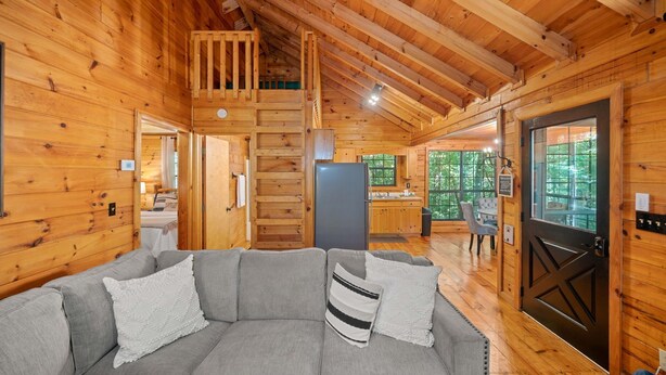 A log cabin interior with a gray sofa, wooden walls, a kitchen area, and a view of the outdoors. - Timber Nest | Stay Amongst the Trees (Blue Ridge)