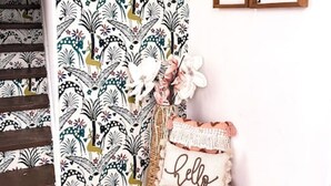 Un escalier orné d’un papier peint à motifs, un banc agrémenté d’un coussin sur lequel est écrit « bonjour », et des œuvres d’art encadrées accrochées au mur.