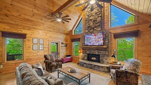 A log cabin interior with a stone fireplace, wooden ceiling, and large windows. - Mountain Bliss Chalet (Blue Ridge)