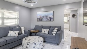 Een moderne woonkamer met grijze banken, een rond poef met nautische patronen, een houten bijzettafeltje en een groot schilderij met een strandthema aan de muur.
